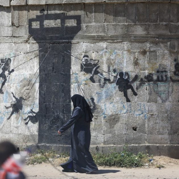 Palestinian women walk past graffiti by British artist Banksy as mounds of rubble and destroyed buildings continue to scar the landscape of Gaza on April 2, 2016. The devastation across Gaza can still be seen nearly two year on from the 2014 conflict between Israel and Palestinian militants. (Photo by Momen Faiz/NurPhoto) (Photo by Momen Faiz / NurPhoto / NurPhoto via AFP)