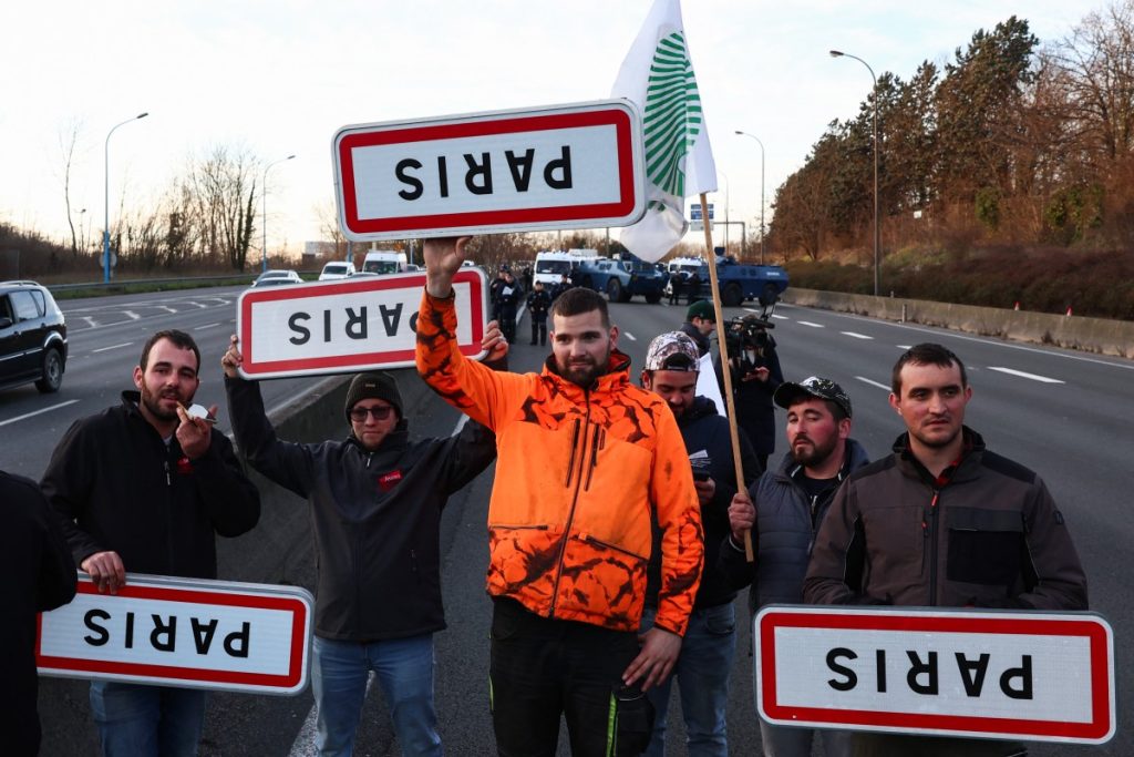 French farmers hold official signs, upside down, of Paris as they block the A6 highway near Chilly-Mazarin, south of Paris, on January 31, 2024 as French farmers maintain roadblocks on key highways into Paris for a third day, as part of nationwide protests called by several farmers' unions over pay, tax and regulations. - Convoys of tractors edged closer to Paris, Lyon and other strategic locations in France on January 31, as thousands of protesting farmers appeared to ignore warnings of police intervention if they cross red lines laid down by the government. Farmers' unions, unimpressed by concessions offered by President Emmanuel Macron's government, encouraged their members to fight on for improved pay, less red tape and protection from foreign competition. (Photo by Emmanuel Dunand / AFP)