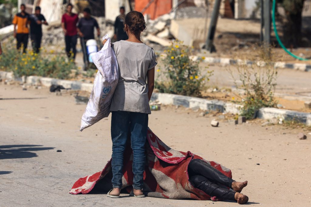 A Palestinian girl stands over the covered body of a person who was killed while seeking food at a distribution point run by the US and Israeli-backed Gaza Humanitarian Foundation (GHF) group, on Salah al-Din road in Nusseirat in the central Gaza Strip on August 4, 2025. Aid agencies have warned that Gaza's population is facing a catastrophic famine, triggered by Israeli restrictions on aid. (Photo by AFP)