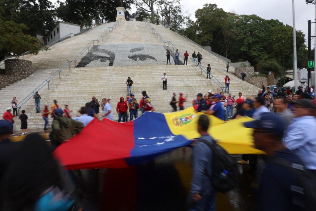 Demonstrators hold a large Venezuelan national flag as they pass by a mosaic portraying the eyes of late Venezuelan President Hugo Chavez on the stairs at Calvario Square during a march in support of ousted Venezuelan President Nicolas Maduro, called by workers in Caracas on January 14, 2026. Venezuela's interim president Delcy Rodriguez declared that her country was entering a new era marked by greater tolerance towards political rivals, following the US ouster of her former boss Nicolas Maduro. (Photo by Pedro MATTEY / AFP)