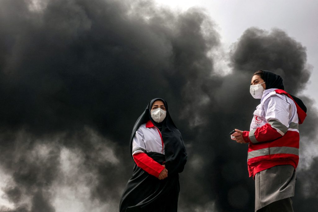 Women members of Iran's Red Crescent society stand near smoke plumes from an ongoing fire following an overnight airstrike on the Shahran oil refinery in northwestern Tehran on March 8, 2026. The United States and Israel launched strikes against Iran on February 28, sparking swift retaliation by the Islamic republic which responded with missile attacks across the region. The war has dragged in global powers, upended the world's energy and transport sectors, and brought chaos to even usually peaceful areas of the volatile region. (Photo by AFP)