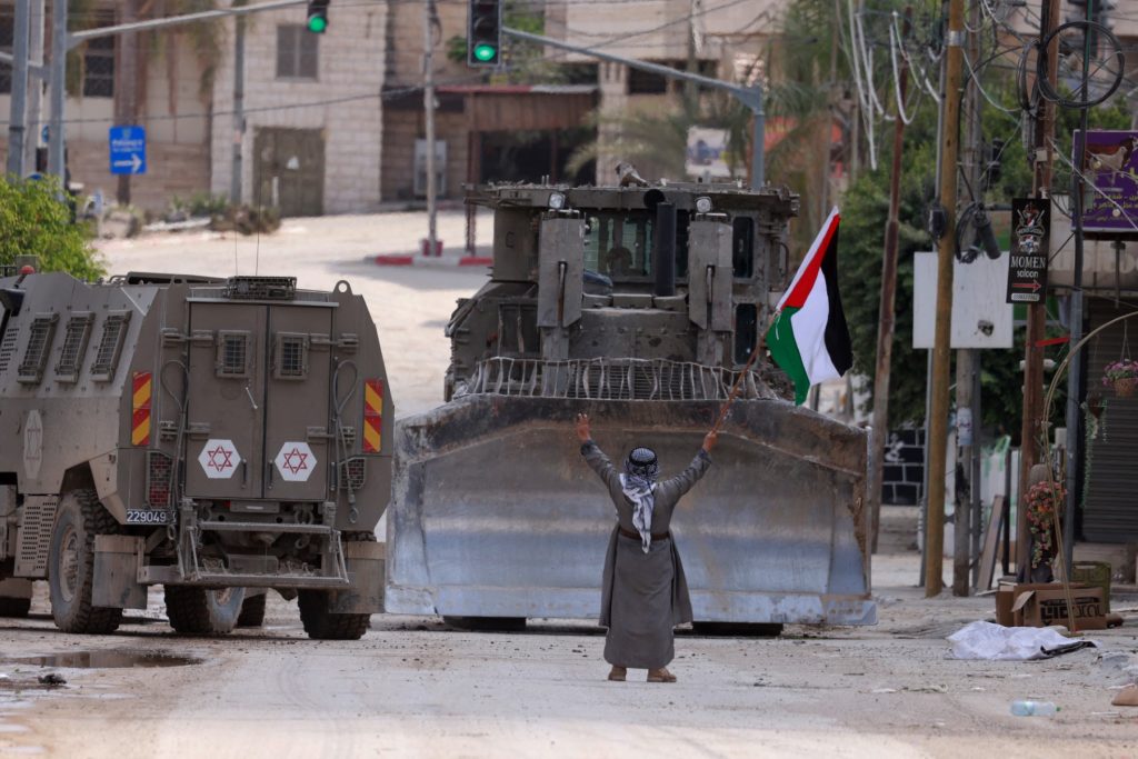 A Palestinian activist lifts a national flag and flashes the victory sign as Israeli armoured vehicles including a bulldozer drive on a street during a raid in Tulkarem on September 3, 2024, amid a large-scale military offensive launched a week earlier in the occupied West Bank. (Photo by Jaafar ASHTIYEH / AFP)
