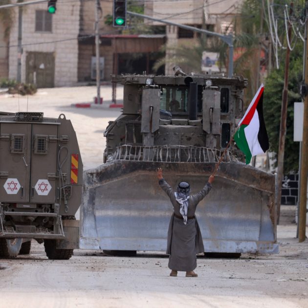 A Palestinian activist lifts a national flag and flashes the victory sign as Israeli armoured vehicles including a bulldozer drive on a street during a raid in Tulkarem on September 3, 2024, amid a large-scale military offensive launched a week earlier in the occupied West Bank. (Photo by Jaafar ASHTIYEH / AFP)