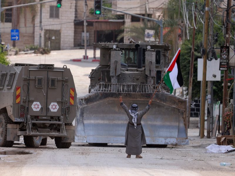 A Palestinian activist lifts a national flag and flashes the victory sign as Israeli armoured vehicles including a bulldozer drive on a street during a raid in Tulkarem on September 3, 2024, amid a large-scale military offensive launched a week earlier in the occupied West Bank. (Photo by Jaafar ASHTIYEH / AFP)