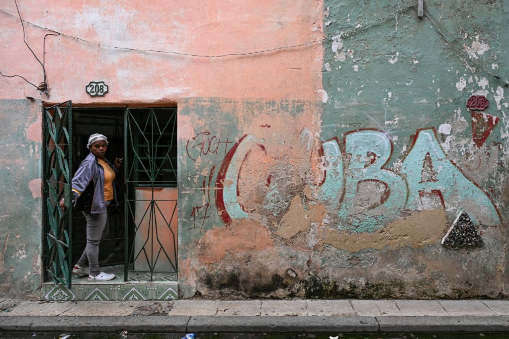 A woman peeks out from the doorway of her home next to a wall with a graffiti reading CUBA on a street in Havana, Cuba, on May 6, 2026. Havana accused Marco Rubio of "lying" after the US Secretary of State stated on May 6, 2026 that Washington had not imposed an oil embargo on Cuba and attributed the island's current energy crisis to internal mismanagement. (Photo by YAMIL LAGE / AFP)