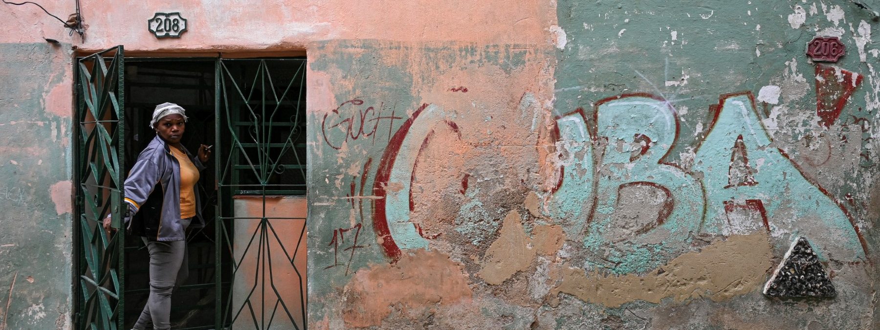 A woman peeks out from the doorway of her home next to a wall with a graffiti reading CUBA on a street in Havana, Cuba, on May 6, 2026. Havana accused Marco Rubio of "lying" after the US Secretary of State stated on May 6, 2026 that Washington had not imposed an oil embargo on Cuba and attributed the island's current energy crisis to internal mismanagement. (Photo by YAMIL LAGE / AFP)