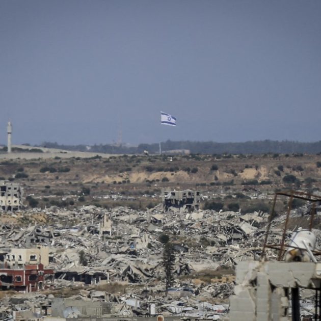 A view shows destroyed buildings and an Israeli flag visible in the distance in northern Gaza on May 6, 2026. Palestinian sisters Aya Zaid and Lina Zaid said they are carrying out a humanitarian initiative nearby to distribute meals to children in an area sheltering displaced families near the yellow line, where food aid remains limited. (Photo by Saeed Jaras / Middle East Images via AFP)