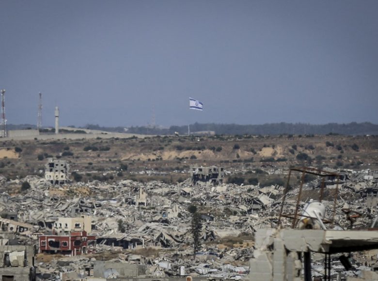 A view shows destroyed buildings and an Israeli flag visible in the distance in northern Gaza on May 6, 2026. Palestinian sisters Aya Zaid and Lina Zaid said they are carrying out a humanitarian initiative nearby to distribute meals to children in an area sheltering displaced families near the yellow line, where food aid remains limited. (Photo by Saeed Jaras / Middle East Images via AFP)