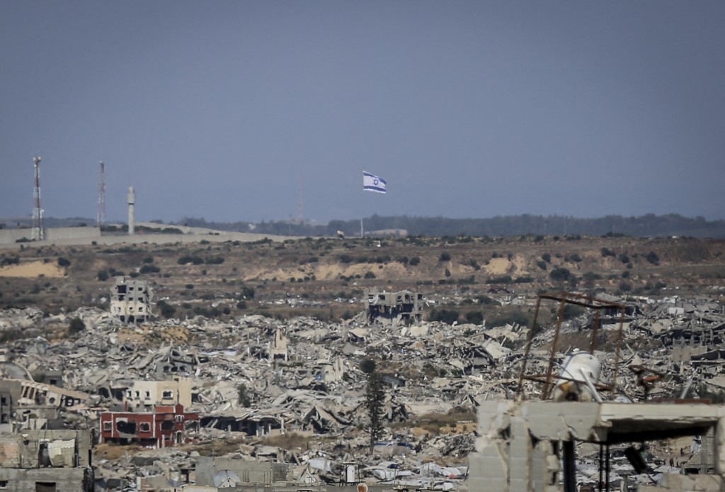 A view shows destroyed buildings and an Israeli flag visible in the distance in northern Gaza on May 6, 2026. Palestinian sisters Aya Zaid and Lina Zaid said they are carrying out a humanitarian initiative nearby to distribute meals to children in an area sheltering displaced families near the yellow line, where food aid remains limited. (Photo by Saeed Jaras / Middle East Images via AFP)