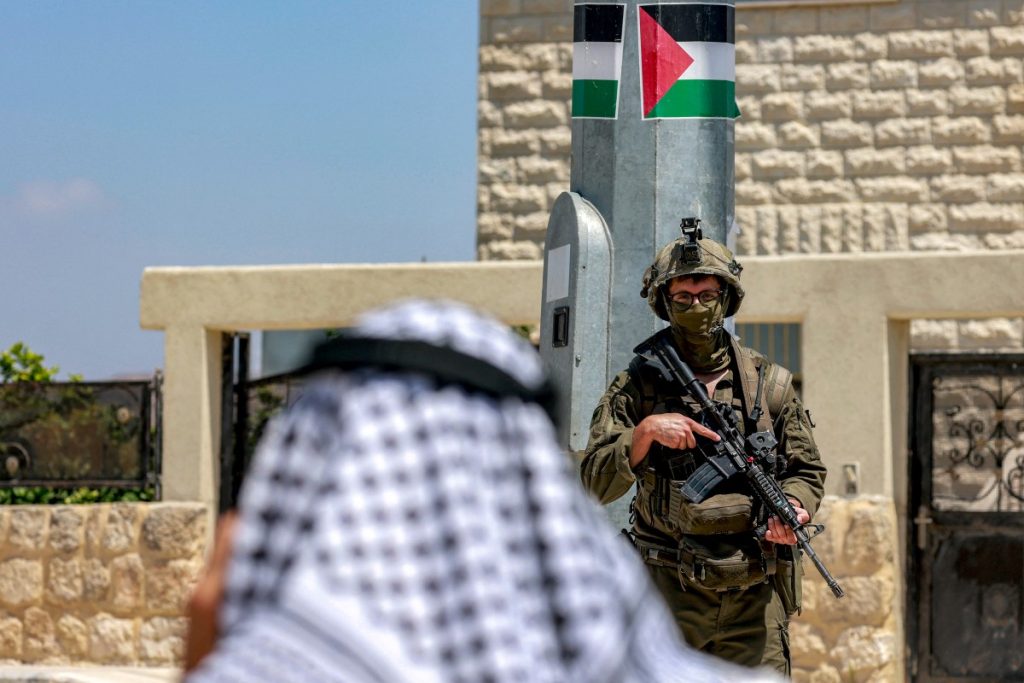A Palestinian man speaks on a phone near Israeli army soldiers standing guard, during an official visit by a group of foreign diplomats accredited by the Palestinian Authority at the archaeological site of Sebastia, north of Nablus in the occupied West Bank, on June 29, 2026. (Photo by Jaafar ASHTIYEH / AFP)