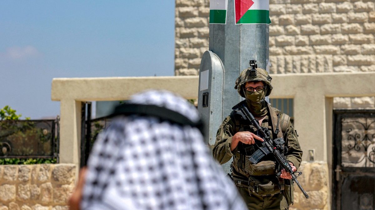 A Palestinian man speaks on a phone near Israeli army soldiers standing guard, during an official visit by a group of foreign diplomats accredited by the Palestinian Authority at the archaeological site of Sebastia, north of Nablus in the occupied West Bank, on June 29, 2026. (Photo by Jaafar ASHTIYEH / AFP)