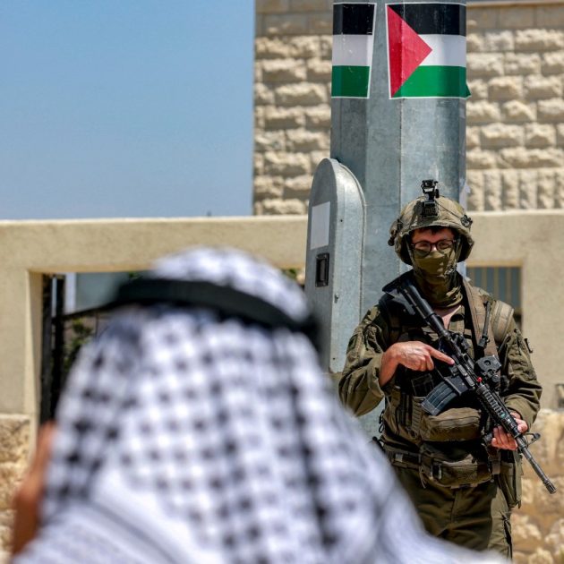 A Palestinian man speaks on a phone near Israeli army soldiers standing guard, during an official visit by a group of foreign diplomats accredited by the Palestinian Authority at the archaeological site of Sebastia, north of Nablus in the occupied West Bank, on June 29, 2026. (Photo by Jaafar ASHTIYEH / AFP)