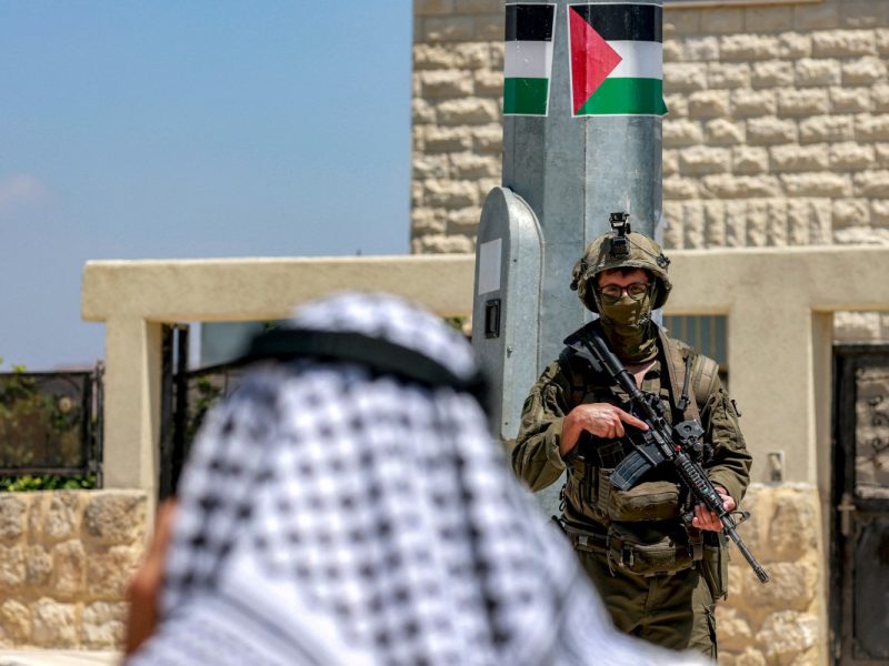 A Palestinian man speaks on a phone near Israeli army soldiers standing guard, during an official visit by a group of foreign diplomats accredited by the Palestinian Authority at the archaeological site of Sebastia, north of Nablus in the occupied West Bank, on June 29, 2026. (Photo by Jaafar ASHTIYEH / AFP)