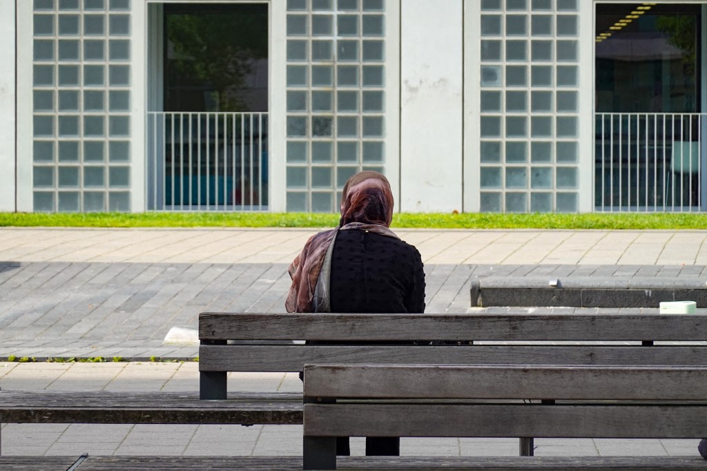 A woman with a headscarf sits alone on a bench, facing a modern building facade in Stuttgart, Baden-Wuerttemberg, Germany, on June 21, 2025. (Photo by Michael Nguyen/NurPhoto) (Photo by Michael Nguyen / NurPhoto via AFP)